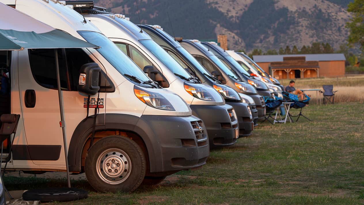Photo of a row of vans lined up. They are parked on grass with mountains in the background